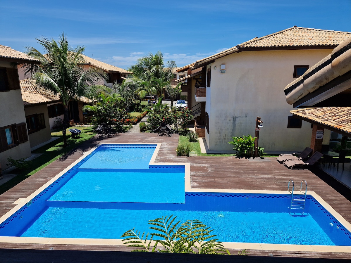 A serene pool area is framed by lush greenery and wooden decking, showcasing two connected pools. Sun loungers are placed beside the pools, and surrounding the area, palm trees and well-maintained gardens create a relaxed atmosphere within the condominium.
