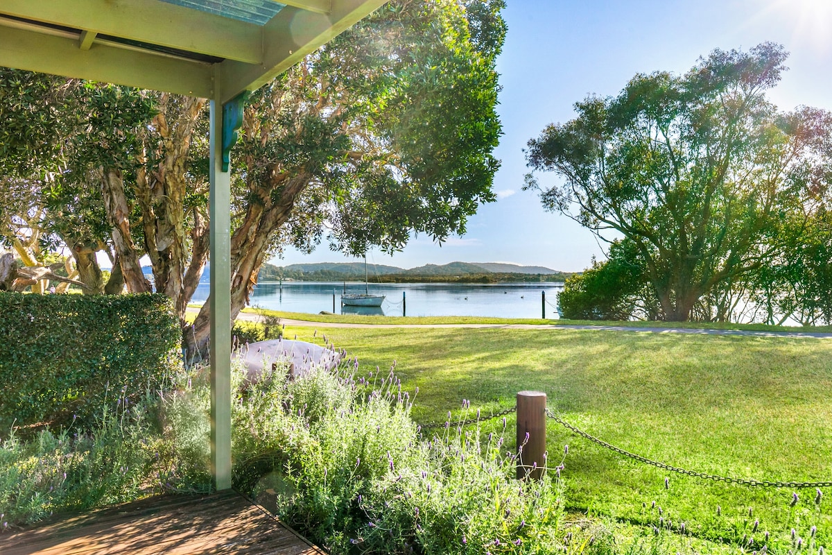 A serene waterside view is captured from the covered deck, showcasing lush green lawns and a backdrop of trees. The tranquil water is visible in the distance, with a boat moored along the shore. Sunlight filters through the foliage, enhancing the peaceful atmosphere.