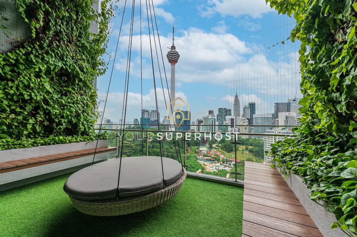 A swing chair positioned on a balcony is surrounded by lush greenery, offering a comfortable place to relax. The panoramic view of Kuala Lumpur's skyline is visible in the background, featuring notable landmarks such as the KL Tower, set against a clear blue sky.