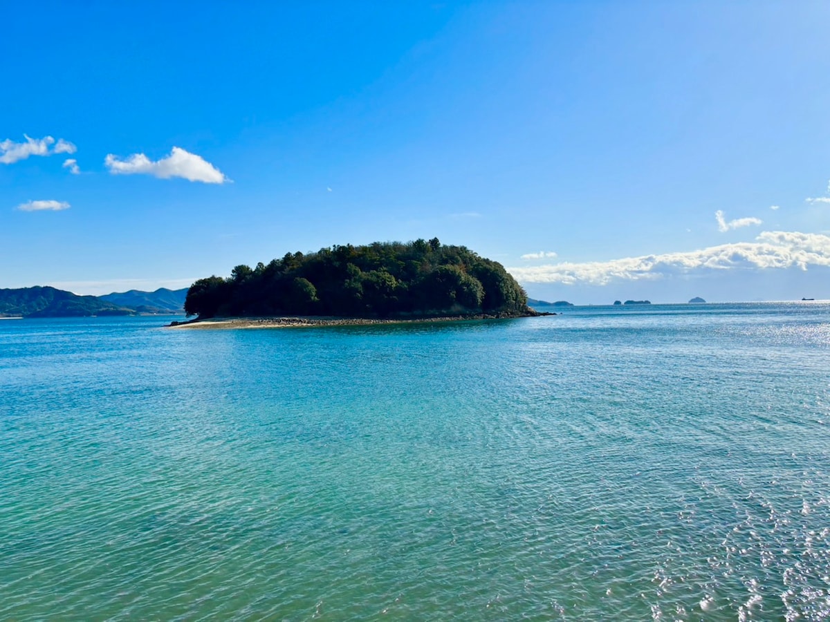 A tranquil island is surrounded by clear turquoise waters under a bright blue sky. Gentle waves can be seen lapping against the shore, while lush greenery covers the island, contributing to the serene coastal landscape.