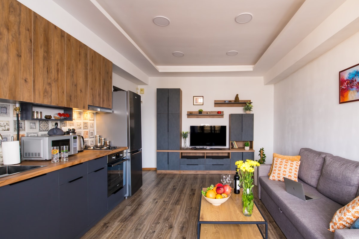 The open-plan living area includes a modern kitchen with a spacious countertop and stainless steel appliances. A comfortable gray sofa is paired with a wooden coffee table, adorned with fresh fruit and drinks. Decorative shelving enhances the ambiance, and a flat-screen TV is prominently displayed.