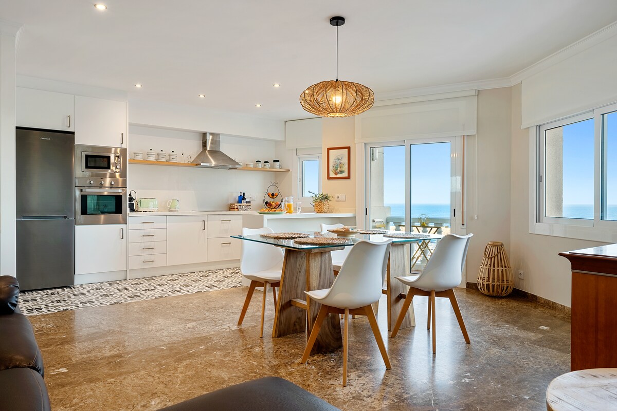An open-concept kitchen and dining area features modern white cabinetry and a large circular wooden table surrounded by comfortable white chairs. Large windows allow natural light to illuminate the space, offering views of the Mediterranean Sea. A stylish pendant light hangs above the dining table.