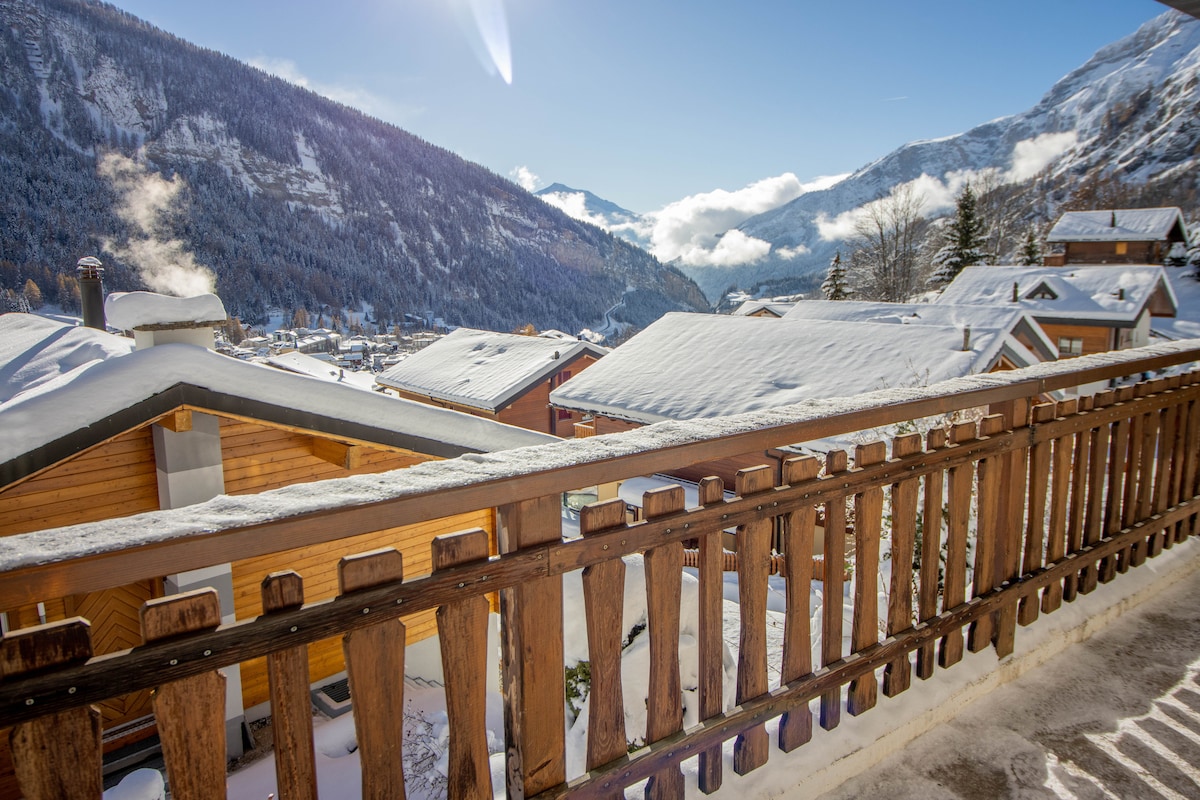 A wooden balcony railing frames a panoramic view of snow-covered rooftops and surrounding mountains. The clear blue sky and soft clouds enhance the serene winter landscape, creating a peaceful atmosphere ideal for reflection.
