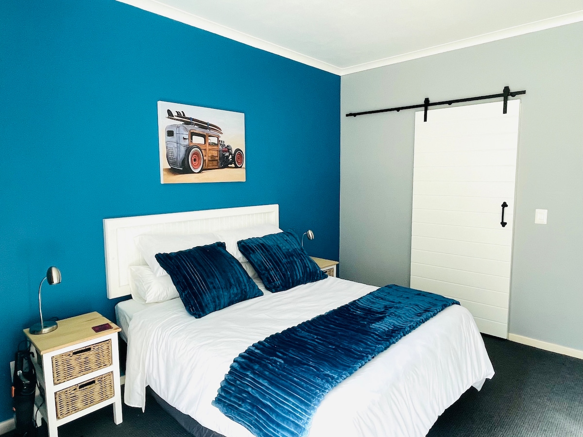A spacious bedroom features a queen-sized bed with a blue bedspread and two coordinated cushions. A striking blue accent wall contrasts with the neutral gray wall. Nightstands with lamps flank the bed, and a sliding barn door adds a decorative touch.