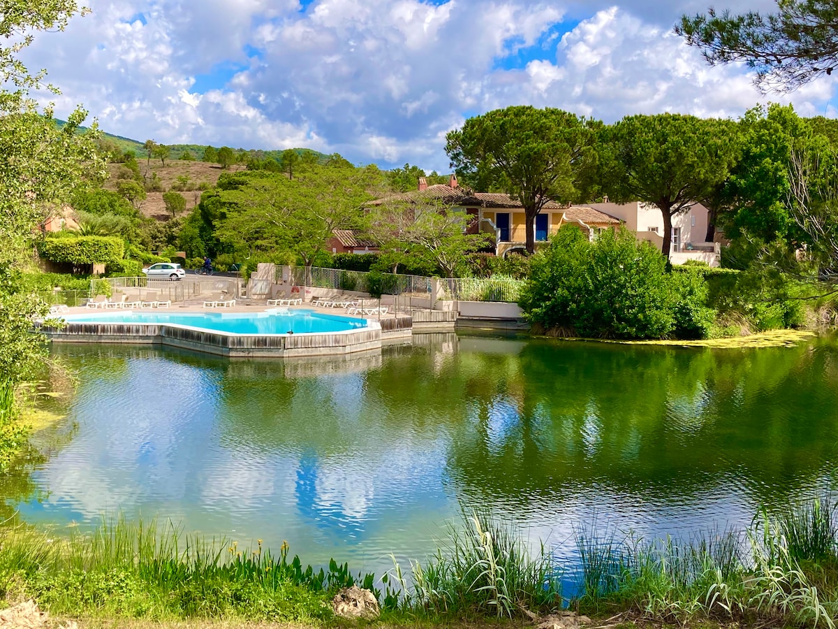 A serene scene showcases a calm lake surrounded by lush greenery and trees. A deck features a pool with a gentle curve, offering a space for relaxation under a partly cloudy sky. Reflections of the landscape are visible in the clear water.
