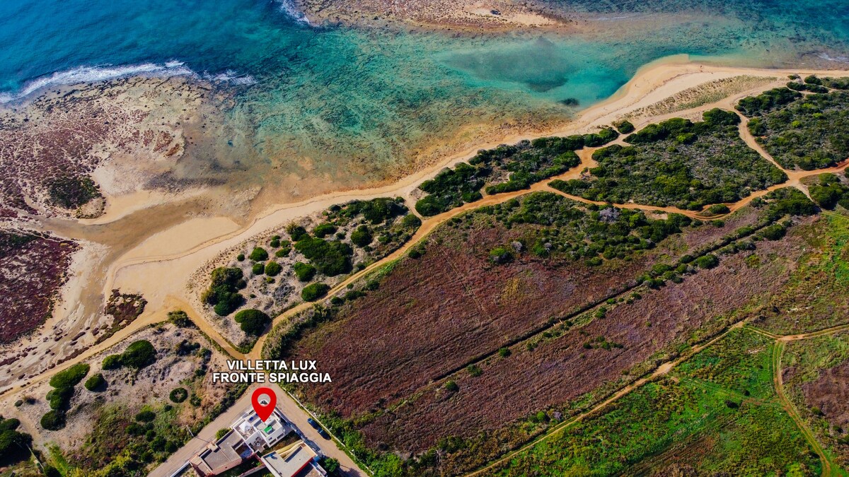 An aerial view captures the coastline of Torre Pali, featuring the sandy beach bordered by clear blue waters. The surrounding green landscape provides a natural contrast to the beach, with paths leading from the villa to the shoreline.