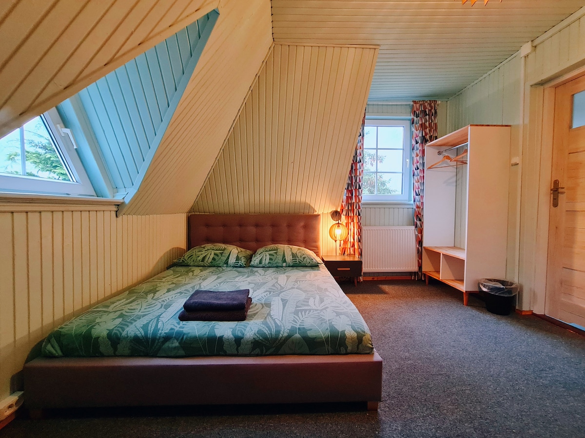 A spacious bedroom features a low-profile bed with a botanical print comforter, complemented by a neatly folded towel. Light enters the room through a window with patterned curtains, while a wardrobe and a small light fixture add to the overall functionality.