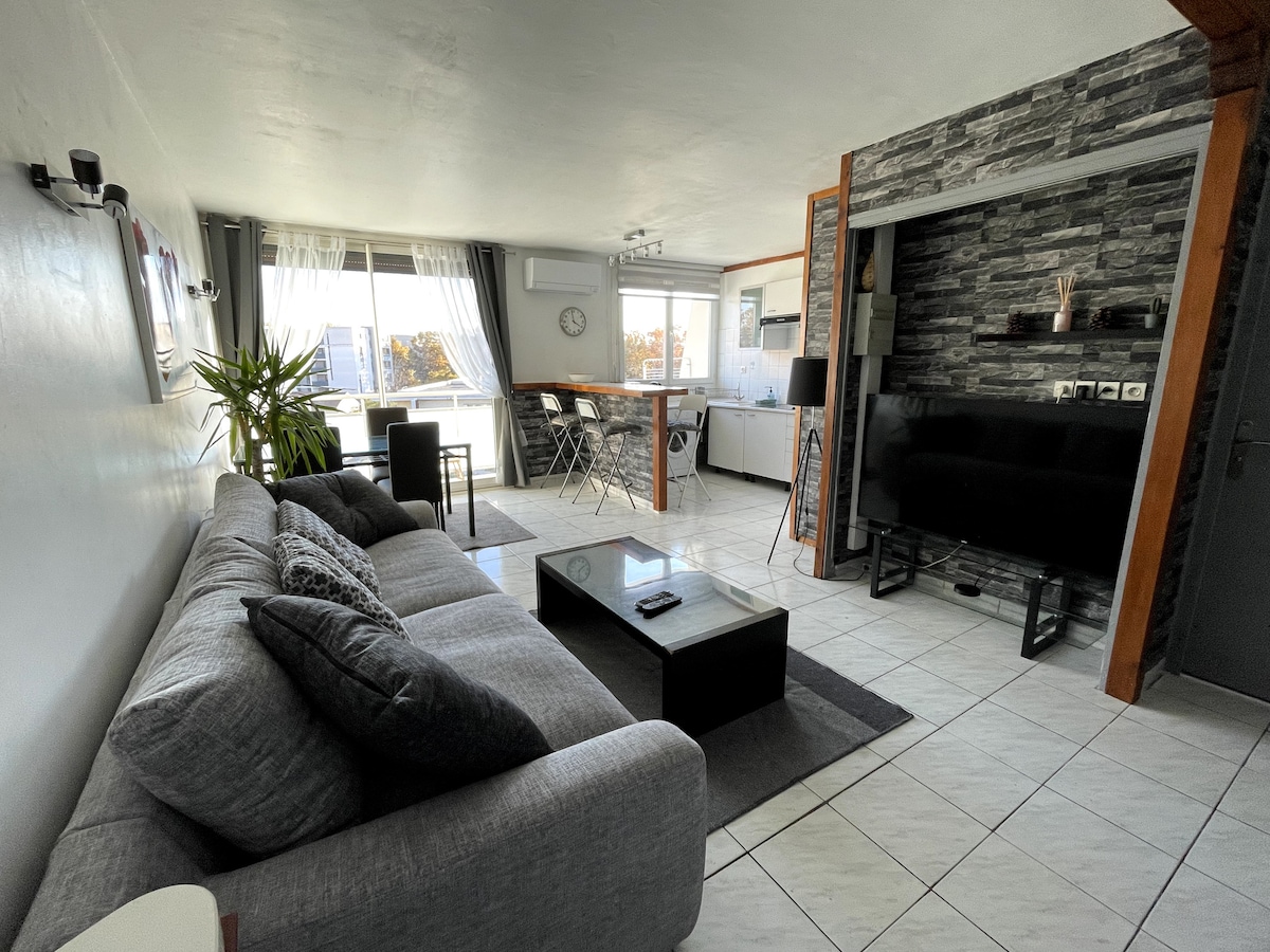 A spacious living area features a comfortable gray sofa complemented by a modern coffee table. Large windows allow ample natural light to fill the room, while the sleek kitchen area is visible in the background, enhancing the open layout. Decorative plants add a touch of greenery.