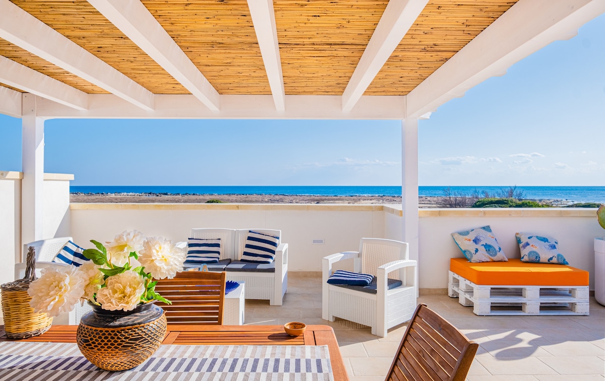 An outdoor terrace is furnished with a wooden dining table and light-colored chairs. A large vase with white flowers is centered on the table. Lounge seating is positioned nearby, with bright orange cushions contrasting the blue ocean view beyond.