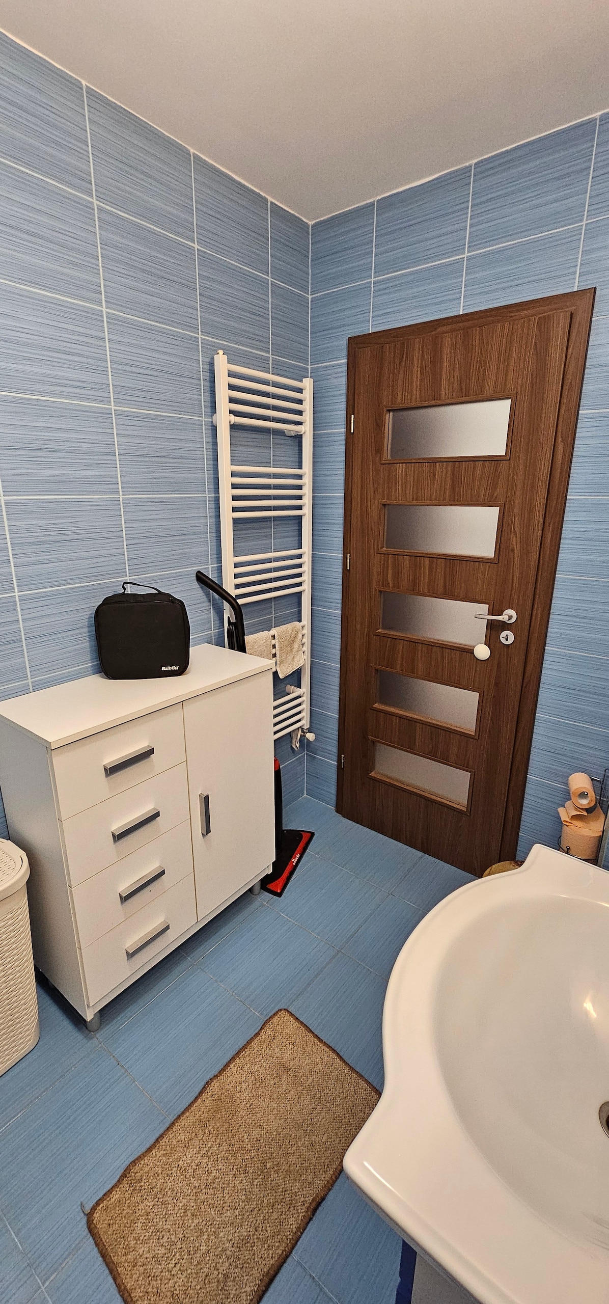 A spacious bathroom features blue tiled walls, with a white shower mat on the floor. A white cabinet with drawers sits next to a sink, while a towel rack is mounted on the wall. A wooden door with frosted glass panels provides access.