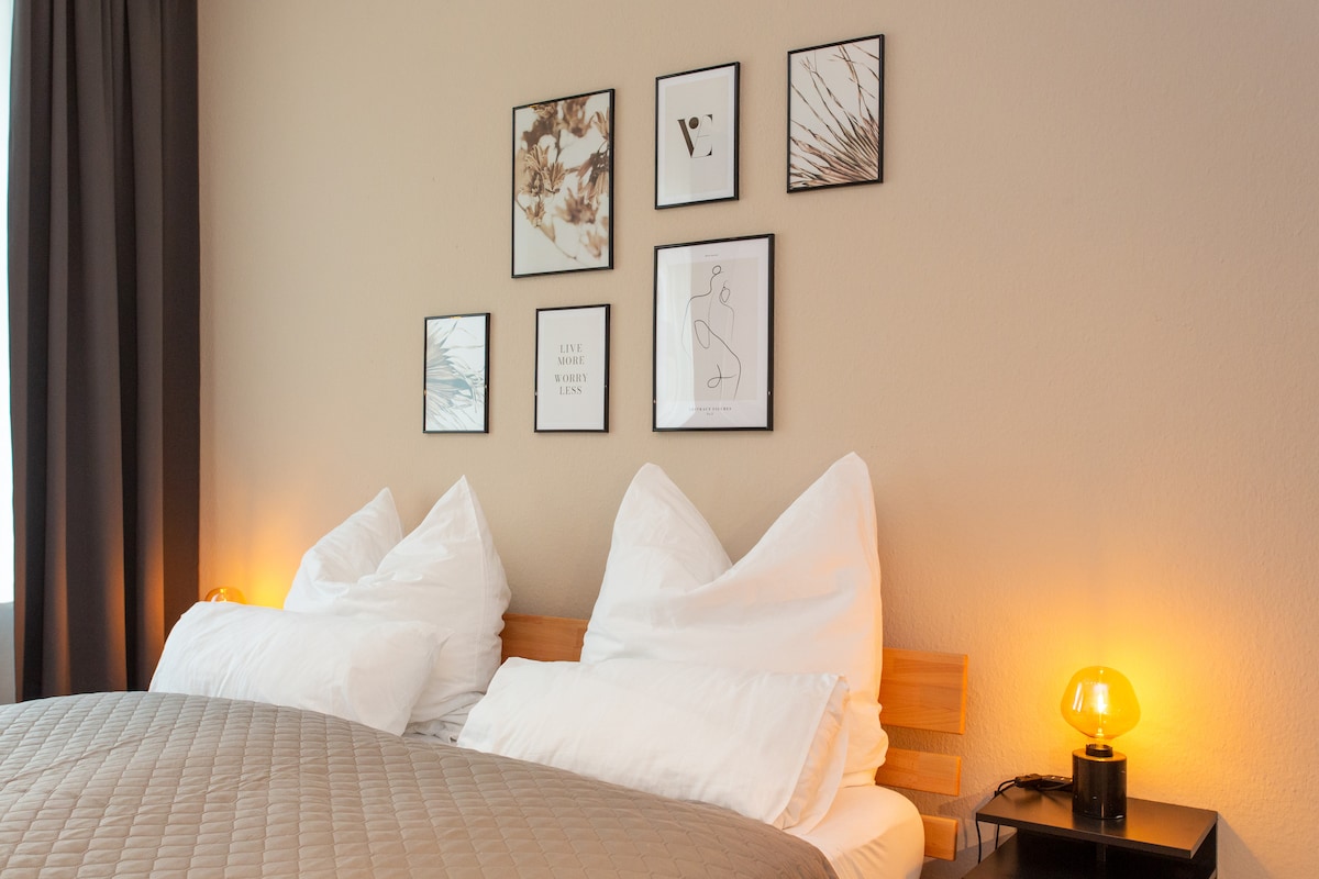 A cozy sleeping area is presented with a wooden bed featuring spacious pillows and crisp white linens. Above the bed, an arrangement of framed art pieces is displayed against a neutral wall. Soft lighting is provided by a stylish bedside lamp.