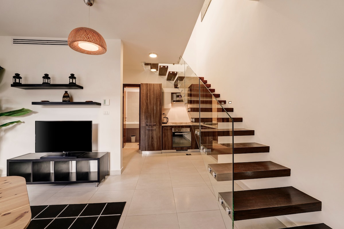 A modern interior features a staircase with wooden steps and a glass railing, leading to an upper level. A sleek black television sits on a contemporary media unit, complemented by a minimalist kitchen visible in the background. Soft lighting contributes to the welcoming ambiance.