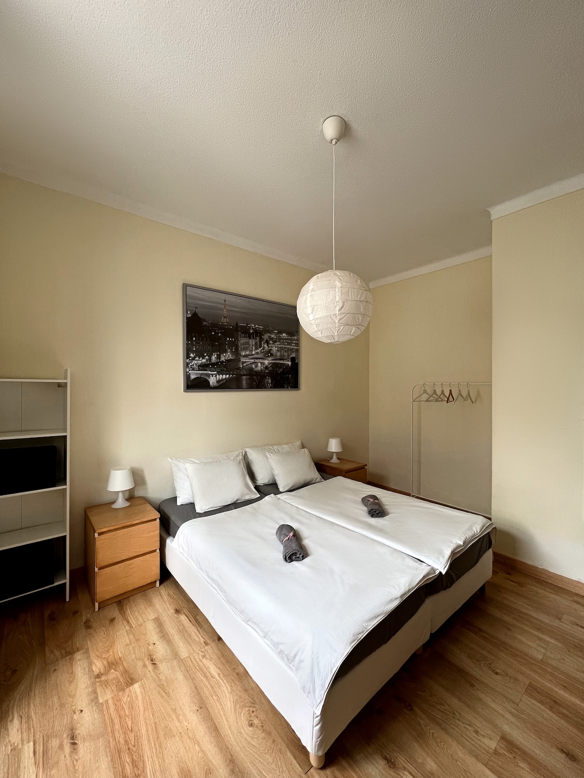 A well-lit bedroom features a spacious bed with white linens and decorative gray accents. Two bedside lamps are placed on wooden nightstands. A black-and-white cityscape photograph hangs above the bed, complemented by a minimalist shelf and a clothing rack in the corner.