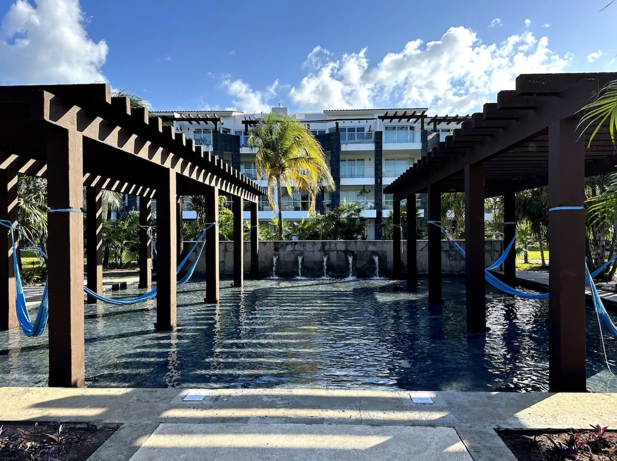 The image features a clear swimming pool situated beneath wooden pergolas, with hanging hammocks visible on either side. A gentle water feature can be observed at the back, while the lush greenery surrounds the pool area, complemented by a building facade in the background.