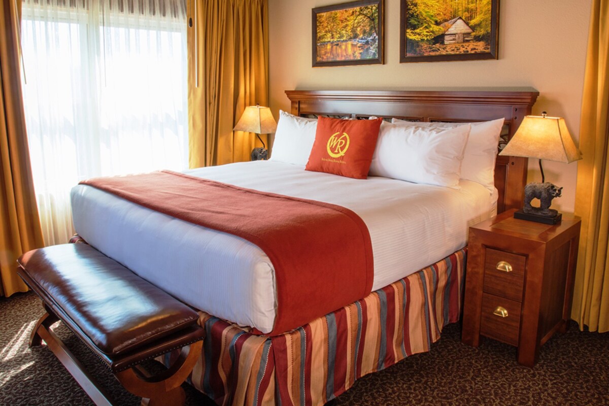 A spacious bedroom is presented with a king bed dressed in crisp white linens, accented by a rich orange throw. Two stylish bedside lamps provide a soft glow, while large windows with light curtains invite natural light into the room. Decorative artwork featuring nature is displayed on the wall.