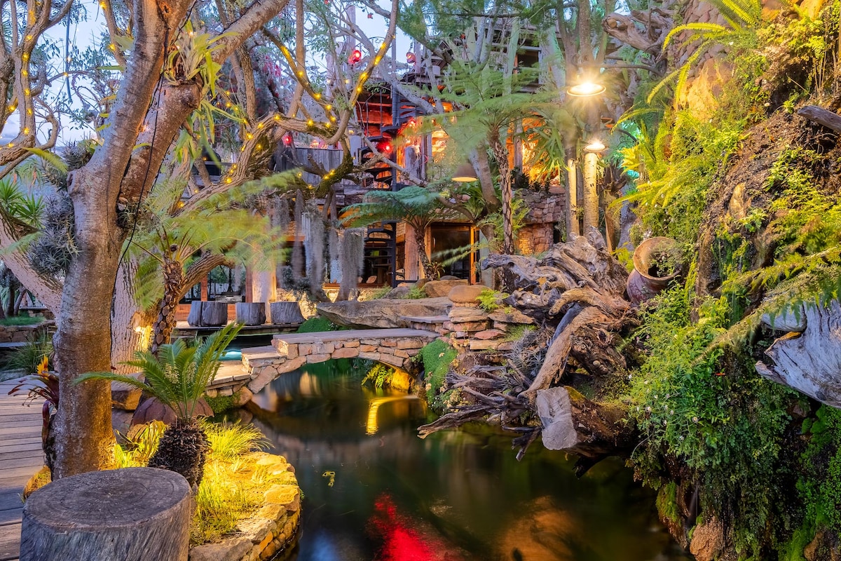 A serene garden area features a winding stone bridge over a natural pool, surrounded by lush vegetation and tall trees. Warm lighting casts a gentle glow on the tranquil water, enhancing the organic beauty of the environment.