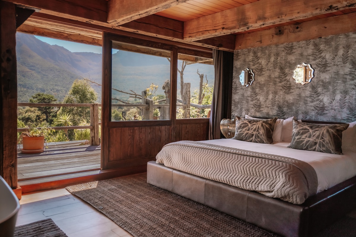 A spacious bedroom features large windows with mountain views, allowing natural light to filter in. The bed is dressed in neutral tones, complemented by decorative pillows. A woven rug covers the floor, and a wooden accent wall enhances the natural aesthetic.