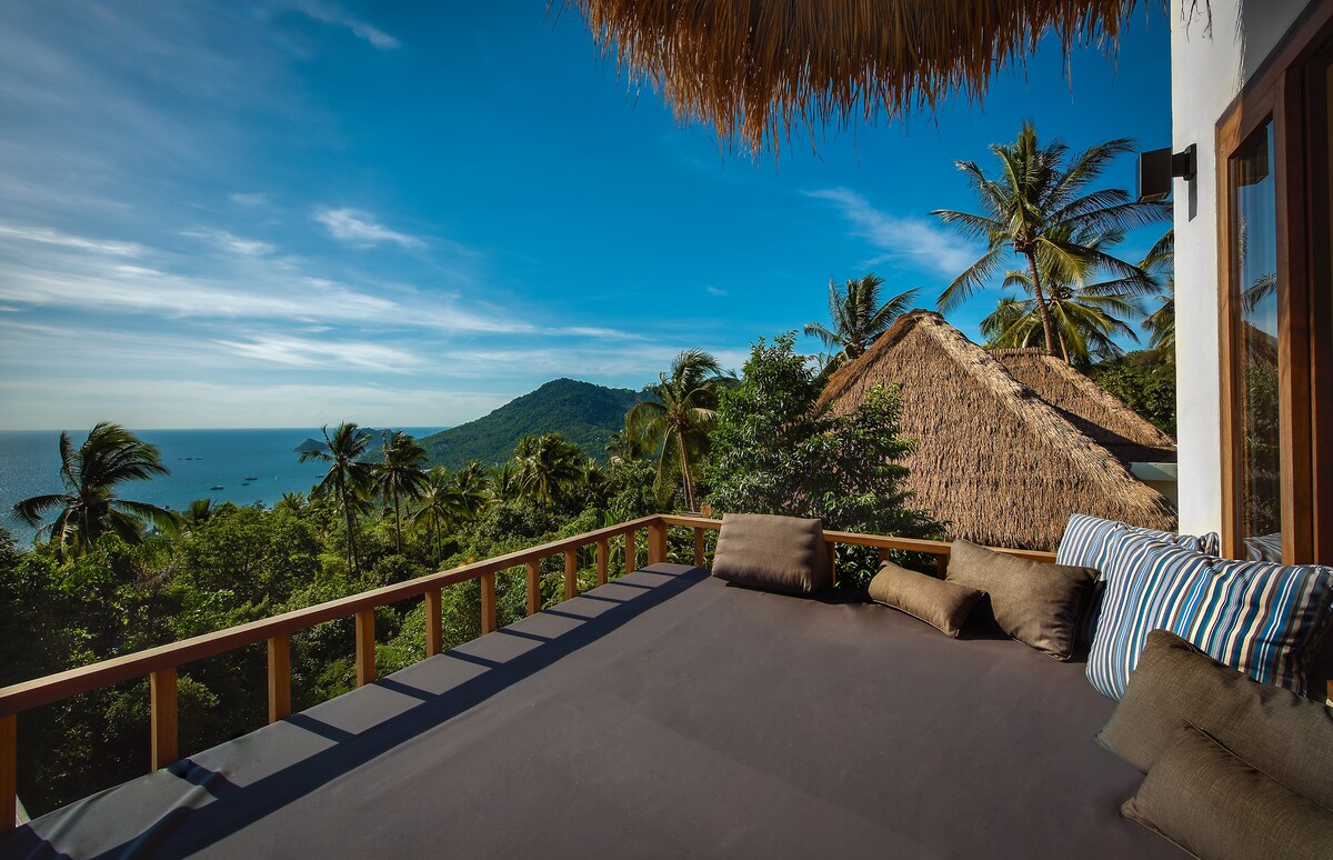 An expansive private sundeck overlooks lush greenery and the scenic coastline. Cushioned seating, featuring earthy tones and striped accents, invites relaxation. The backdrop showcases a tranquil sea view, framed by swaying palm trees and a vibrant blue sky.