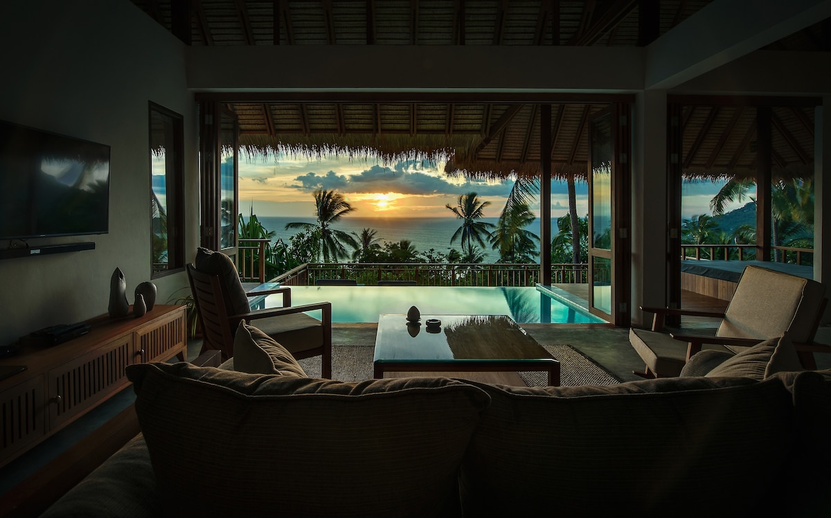 The open living area presents a serene view of the sunset over the sea, framed by tall palm trees. A comfortable sofa faces a wooden coffee table, with an infinity pool visible beyond, creating a seamless connection between indoor and outdoor spaces.