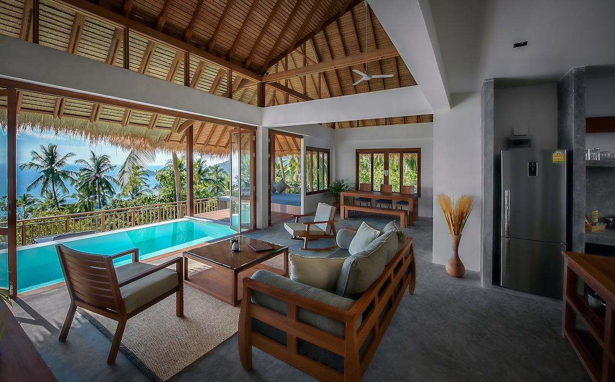 An open living and dining area is showcased with large wooden doors that lead to the outdoor pool. Comfortable seating arrangements in neutral tones are positioned around a wooden coffee table, while large windows offer a view of lush palm trees and the serene pool.