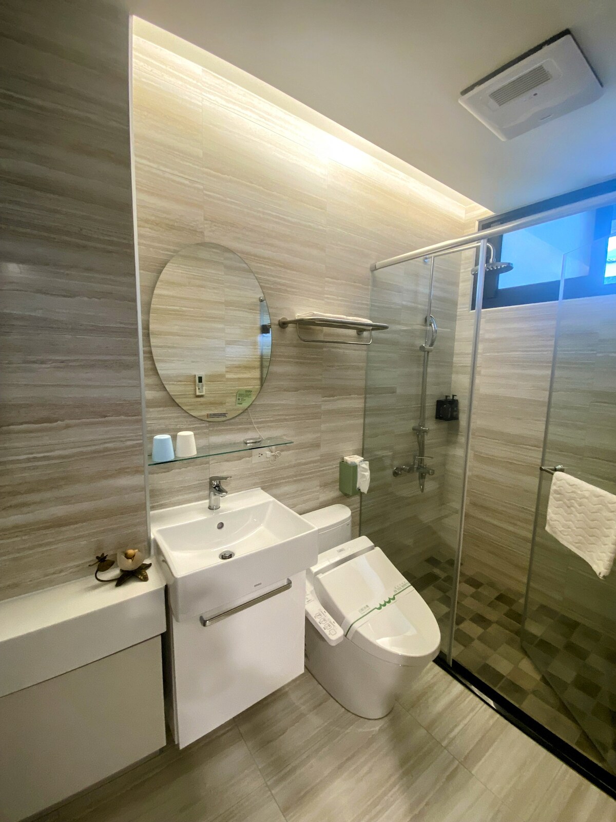 A modern bathroom is presented with a sleek, glass-enclosed shower and minimalist sink. Neutral-toned tiles complement the space, while an oval mirror adds a touch of elegance. Essential toiletries are neatly arranged, and natural light filters in through a small window.
