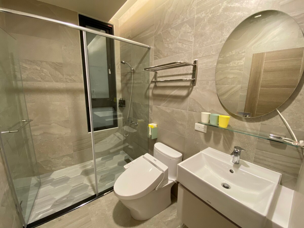 A modern bathroom showcases a glass shower enclosure and a sleek vanity with a white sink. The wall is adorned with light, neutral tiles, and a round mirror reflects the space. Colorful cups sit atop the vanity, adding a subtle touch of warmth.