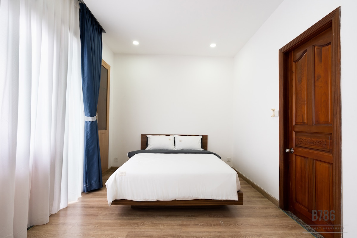 The room features a spacious bed adorned with crisp white linens and a dark throw blanket. Natural light enters through a large window with light curtains, which are complemented by a dark blue curtain. A wooden door adds warmth to the simple design.