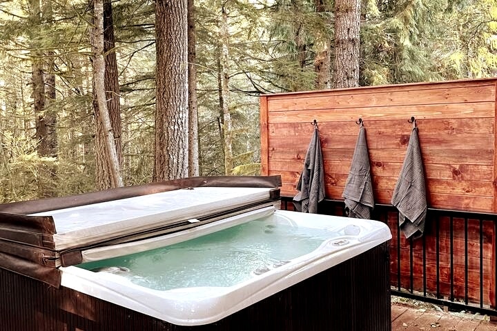 A spacious hot tub is situated on a deck surrounded by tall trees, providing a serene view of the forest. The deck features wooden walls with several towels hanging neatly on hooks nearby.