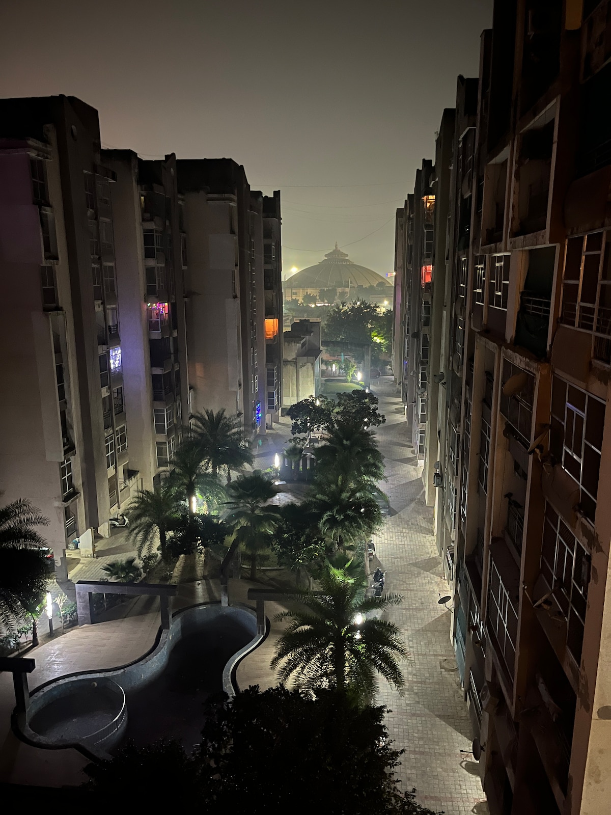 A nighttime view captures a serene pathway lined with palm trees, flanked by modern residential buildings. Soft lights illuminate the walkway, leading toward the distant silhouette of a dome structure, suggesting a nearby temple. A small pool adds a touch of tranquility to the scene.