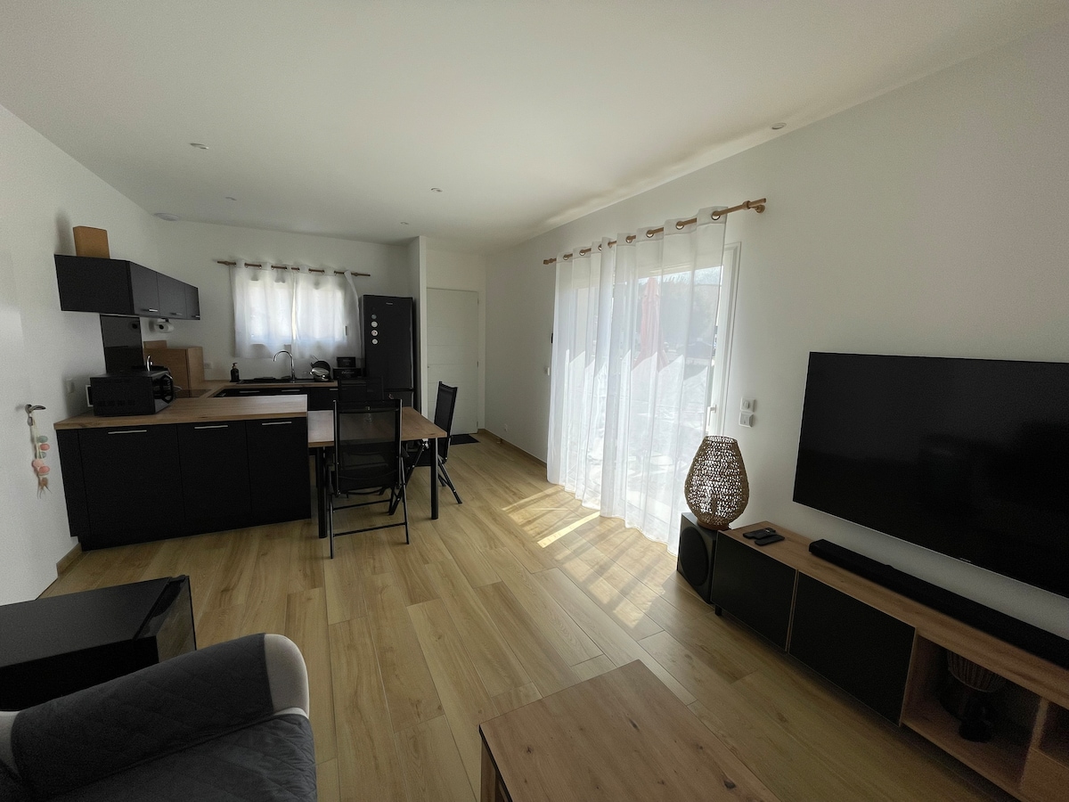 A spacious living and dining area features a modern black kitchen with ample cabinetry, a wooden dining table surrounded by chairs, and a cozy seating arrangement. Large windows allow natural light to fill the space, highlighting the clean lines and neutral decor.