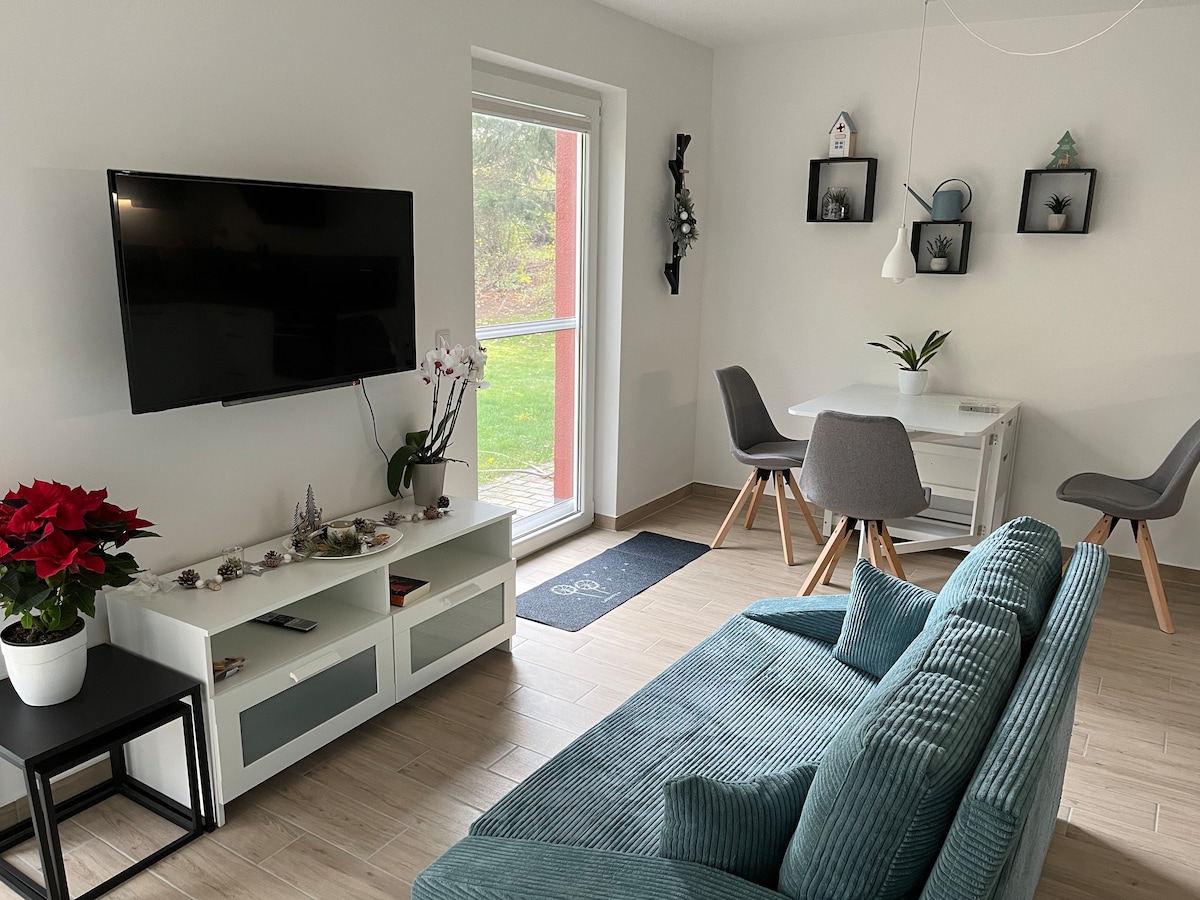 A modern living area displays a comfortable teal sofa and a wall-mounted television. A small dining table with grey chairs is positioned near a window, allowing natural light to illuminate the space. A potted plant adds a touch of greenery to the decor.