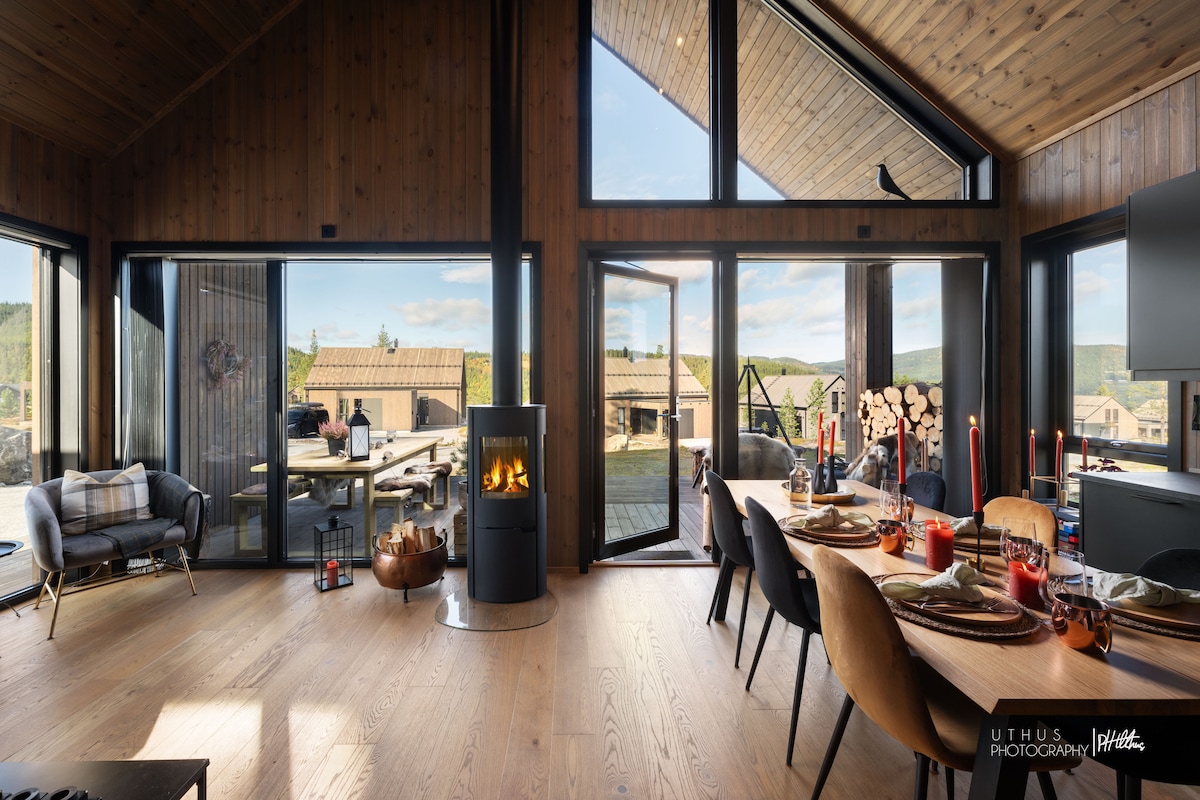 A modern interior is highlighted by wood-paneled walls and large glass doors. A dining table is set with candles, and a cozy seating area includes a stylish armchair. A black fireplace adds warmth, while views of the outdoors are visible through the open doors.