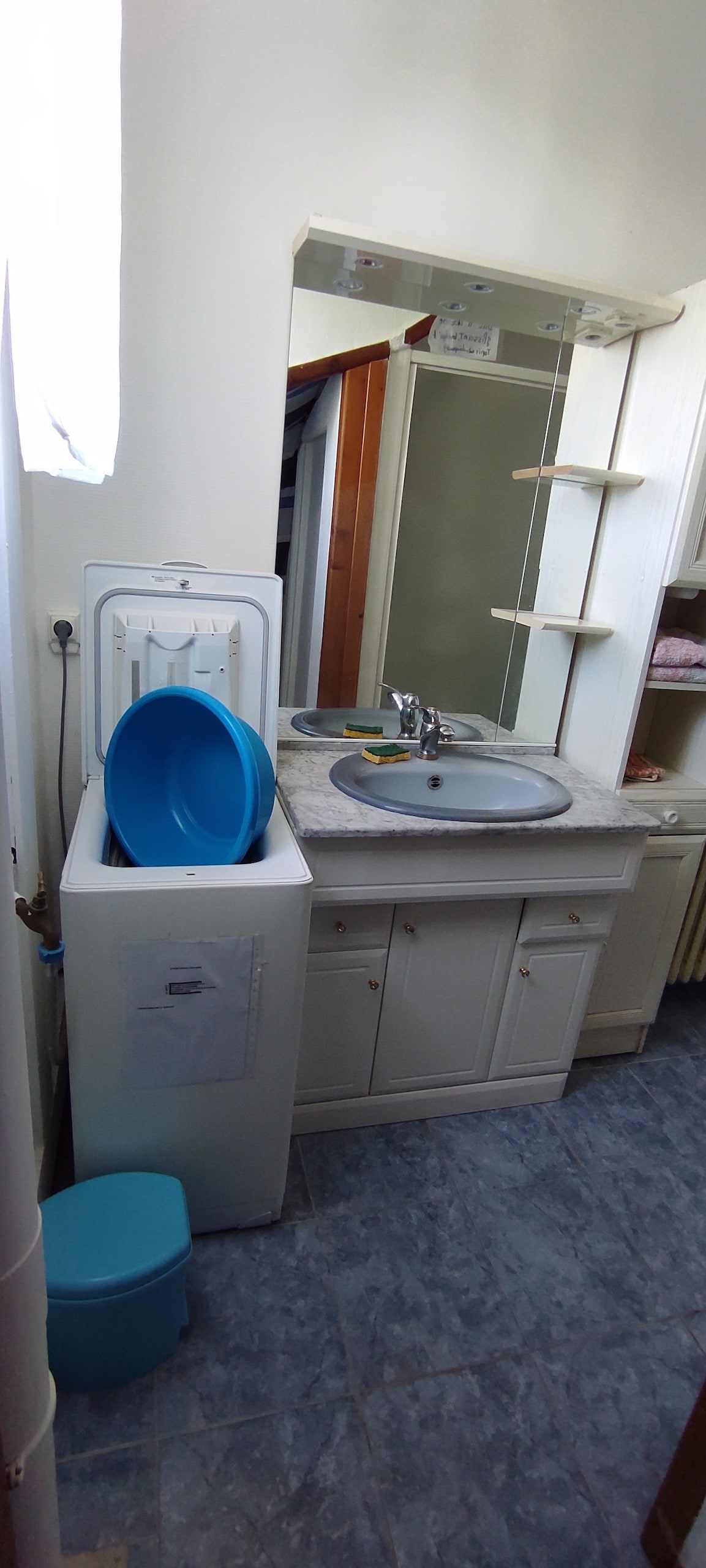 A compact bathroom is shown, featuring a white wash basin and a mirror above it. A washing machine is integrated into the cabinetry, complemented by blue and white containers for storage. Natural light enters through a nearby window, illuminating the tiled flooring.