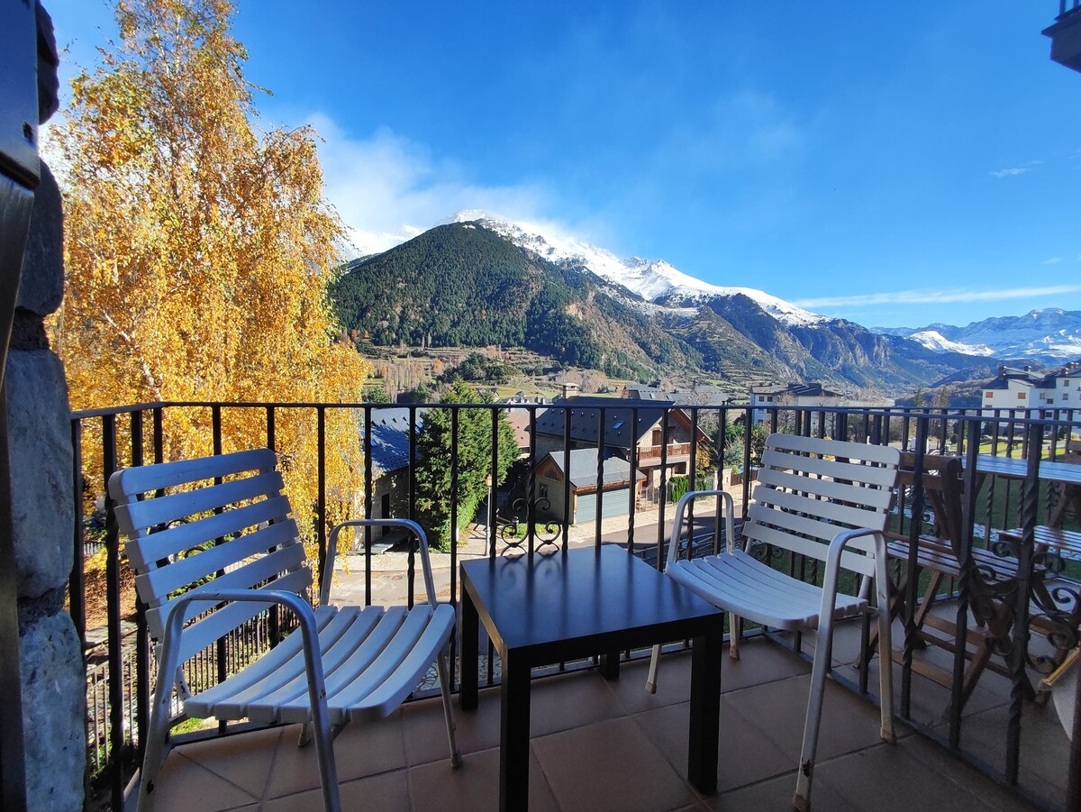 A terrace is showcased with two white chairs and a black table, overlooking a scenic view of mountains. Autumn foliage is visible, and the sky is clear, creating a bright and inviting environment.