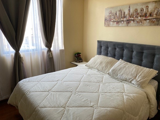 A comfortable sleeping area features a spacious bed with a light-colored quilt. Two pillows are neatly arranged on the bed. Soft natural light enters through large windows, framed by flowing curtains. A small bedside table displays decorative items against a light wall.