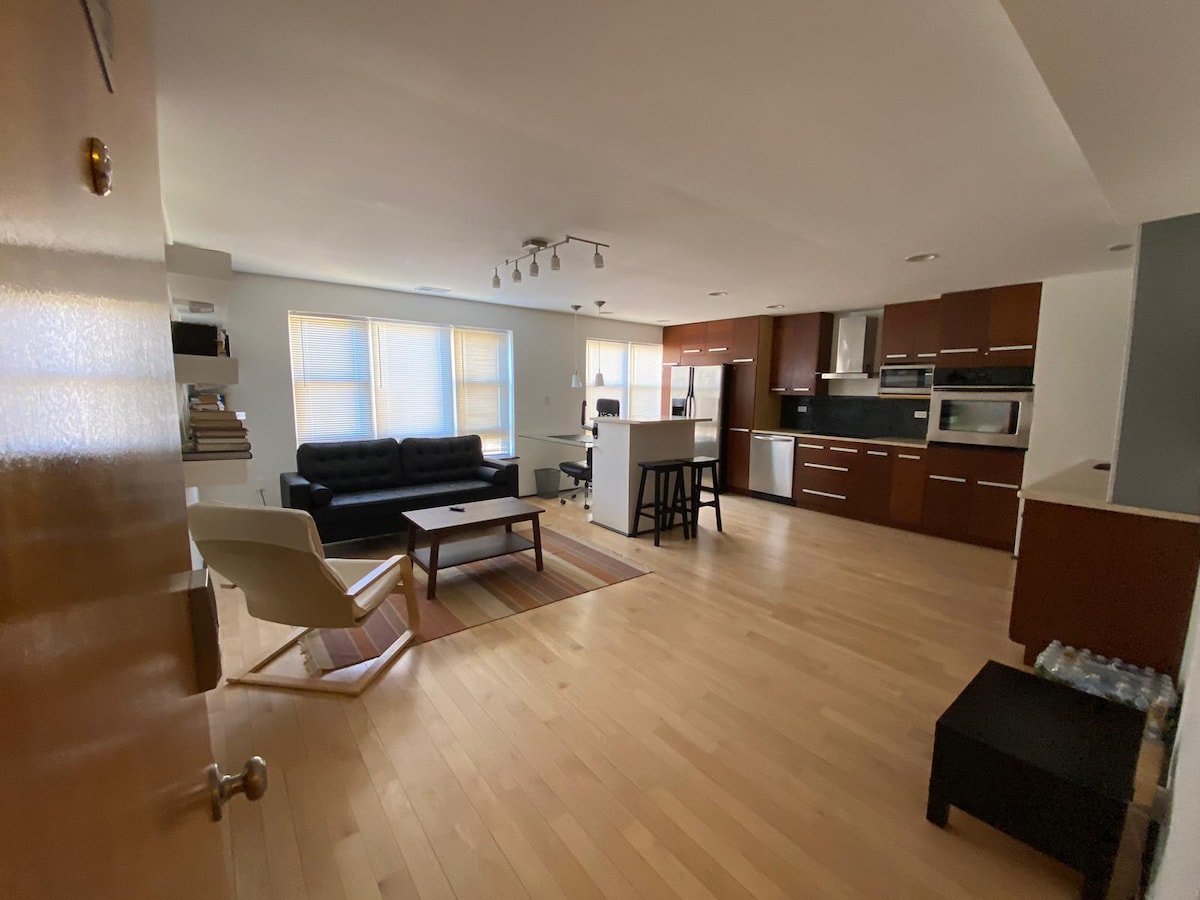 An open-concept living area features hardwood flooring and natural light streaming through large windows. A modern kitchen with dark cabinetry and stainless steel appliances is visible, alongside a comfortable black sofa and a coffee table situated on a patterned rug.