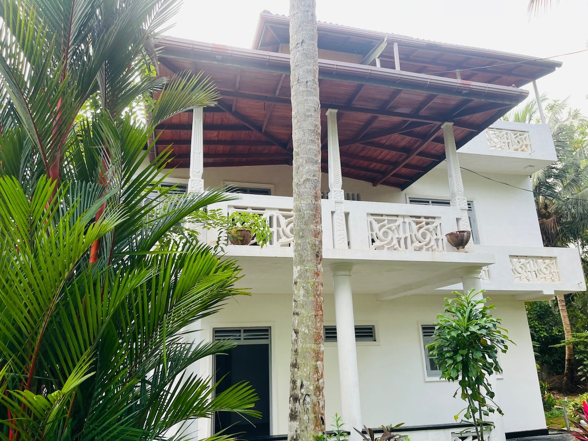 The villa features a two-story structure with a spacious balcony supported by decorative columns. Lush greenery surrounds the building, including palm trees and vibrant plants, creating a natural atmosphere. The white exterior is complemented by traditional design elements, adding character to the overall look.