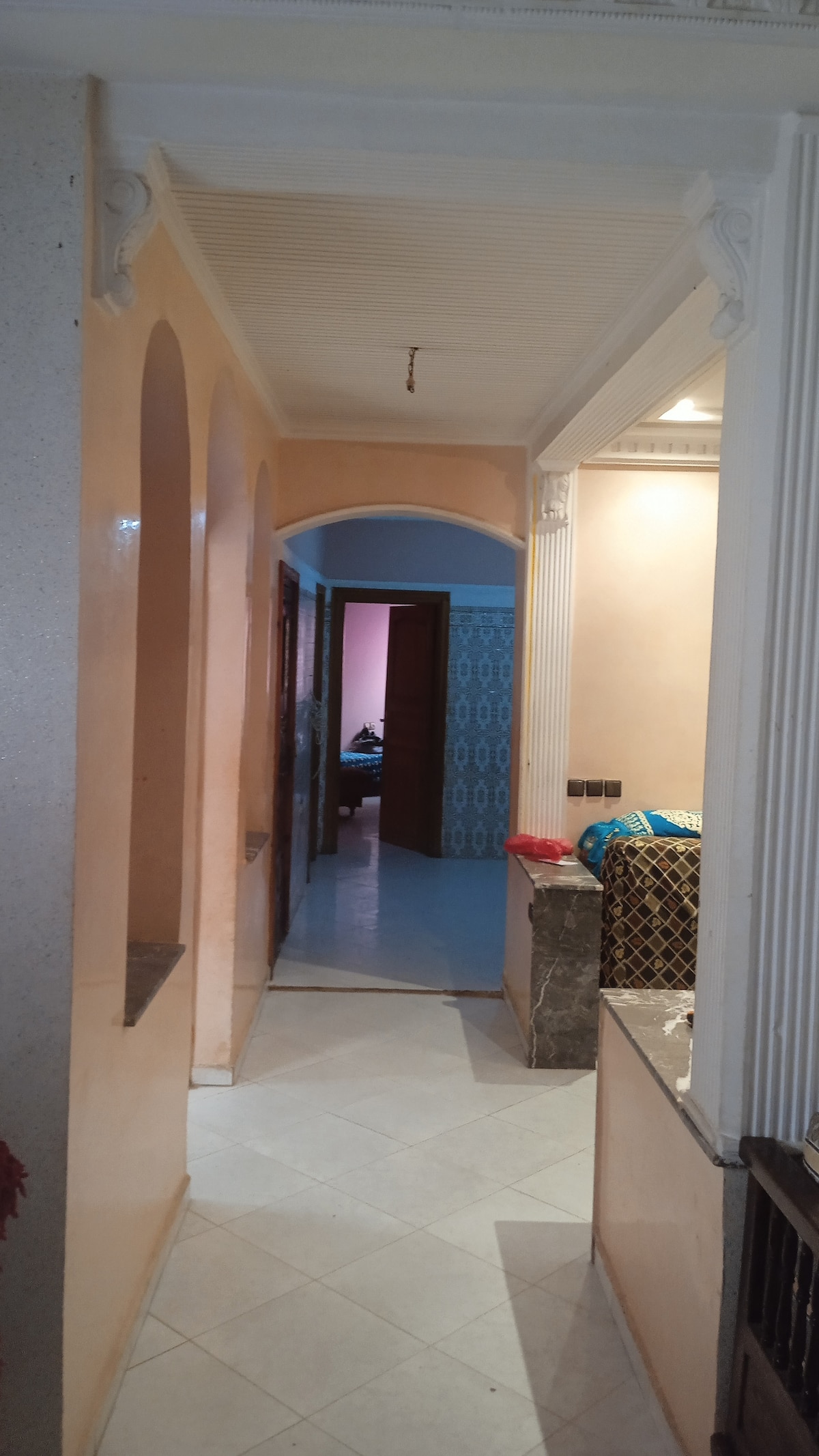 A hallway is presented with soft peach-colored walls and arched doorways that create an airy feel. Light tiles cover the floor, leading toward a partially visible room at the end. Subtle design elements, such as marble accents, enhance the space's character.
