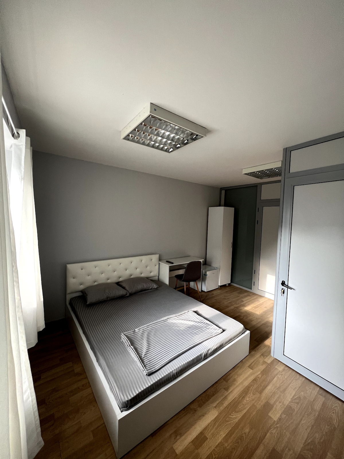 A well-lit bedroom features a spacious bed with a striped grey blanket. A simple desk and chair are positioned near the window, allowing natural light to enhance the neutral color palette. Smooth wooden flooring adds warmth to the overall design.