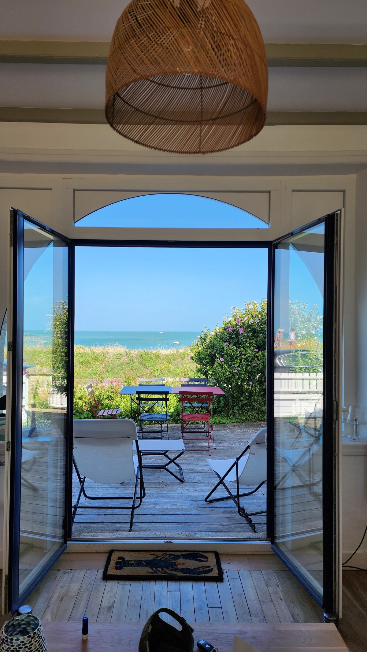 Large glass doors are open, revealing a bright view of the sea and lush greenery outside. A wooden deck is visible, featuring a dining table and chairs set for outdoor enjoyment. Natural light enters the room, enhancing the welcoming environment.