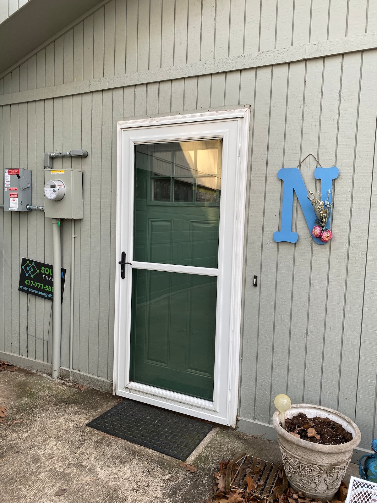 The exterior door showcases a glass panel design with a black handle, leading into the apartment. A decorative blue letter 'N' is displayed beside the door, and an electric meter is visible on the left wall. A small welcome mat lies in front.