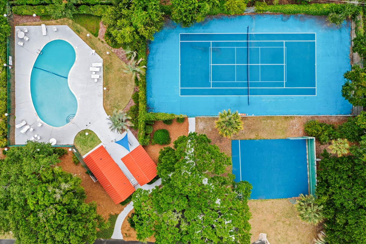 An aerial view displays a private community pool shaped like a kidney, surrounded by lounge chairs and lush greenery. Adjacent to the pool, two tennis courts with bright blue surfaces can be seen, framed by vibrant landscaping.