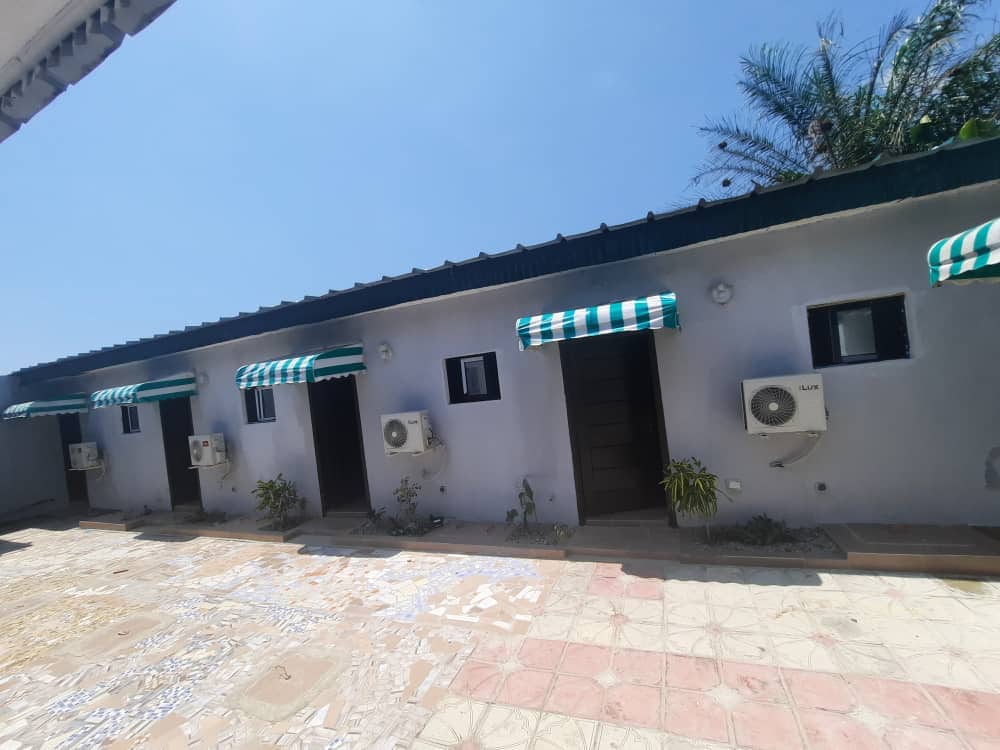 The exterior of the studio building is shown, featuring a pathway leading to individual entrances covered by green and white striped awnings. Air conditioning units are mounted next to each door, and small plants are placed alongside the entrance, set against a clear blue sky.
