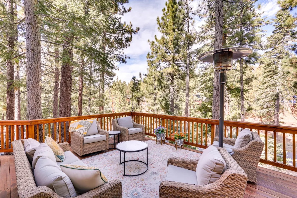 A wraparound deck is surrounded by tall pine trees, with comfortable patio seating arranged around a central coffee table. A gas heater is positioned nearby, and potted plants add a touch of greenery to the serene outdoor space.