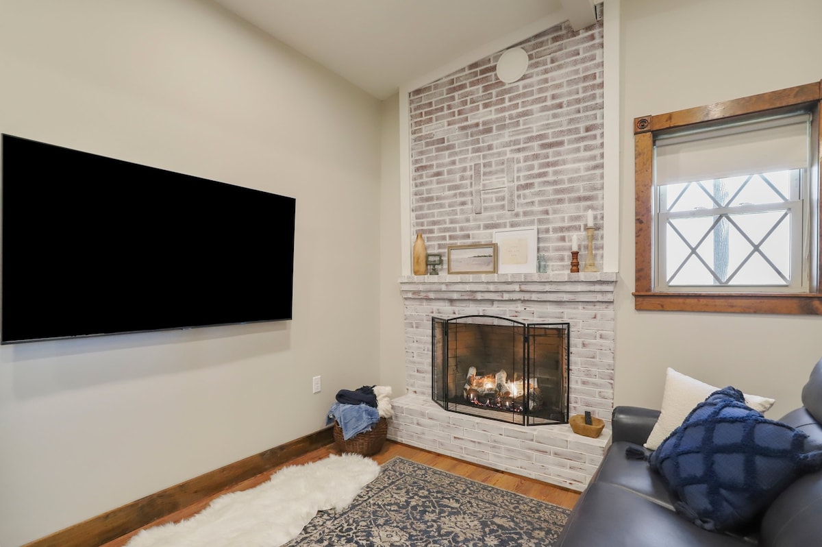 A living room corner features a cozy fireplace built into a white-brick wall, with a sleek flat-screen TV mounted above it. A patterned area rug is placed on the hardwood floor, while a plush throw and decorative accents are arranged nearby, enhancing the relaxing ambiance.