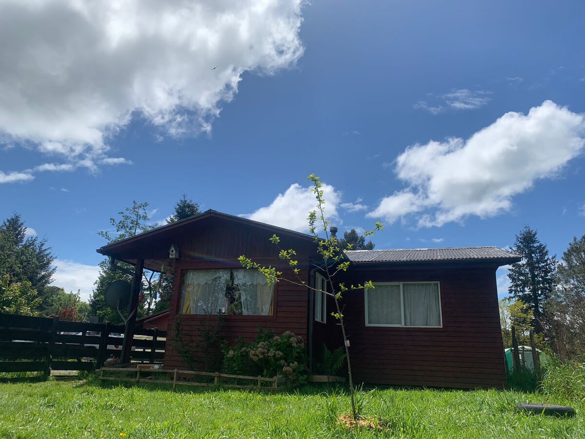 A rustic wooden cabin is framed by lush greenery under a partly cloudy sky. The front porch features a simple railing, while a small tree is visible in the yard, contributing to the tranquil outdoor setting.