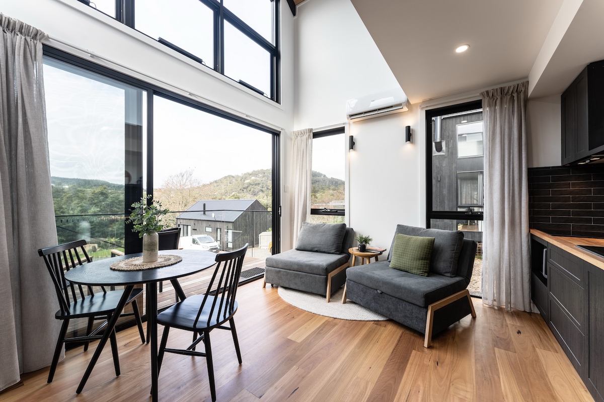 An open living area is highlighted by large windows that provide views of the surrounding landscape. A round dining table with black chairs is positioned near the kitchen, and two gray lounge chairs are arranged for comfort. Warm timber flooring enhances the space.