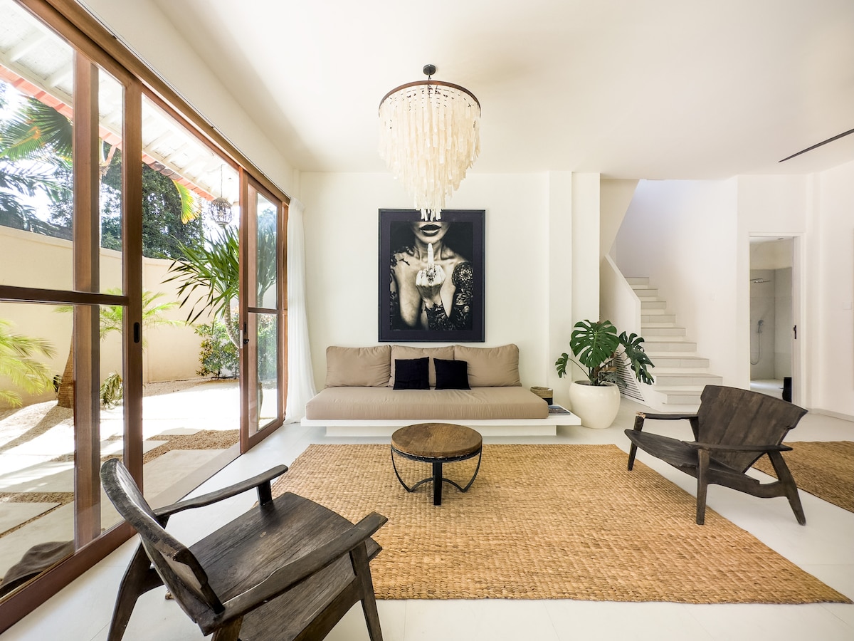 A spacious living area features a large, light-colored sofa, complemented by two wooden chairs and a round coffee table. A striking piece of wall art is mounted above the sofa. Natural light floods the room through expansive glass doors, leading to an outdoor garden.