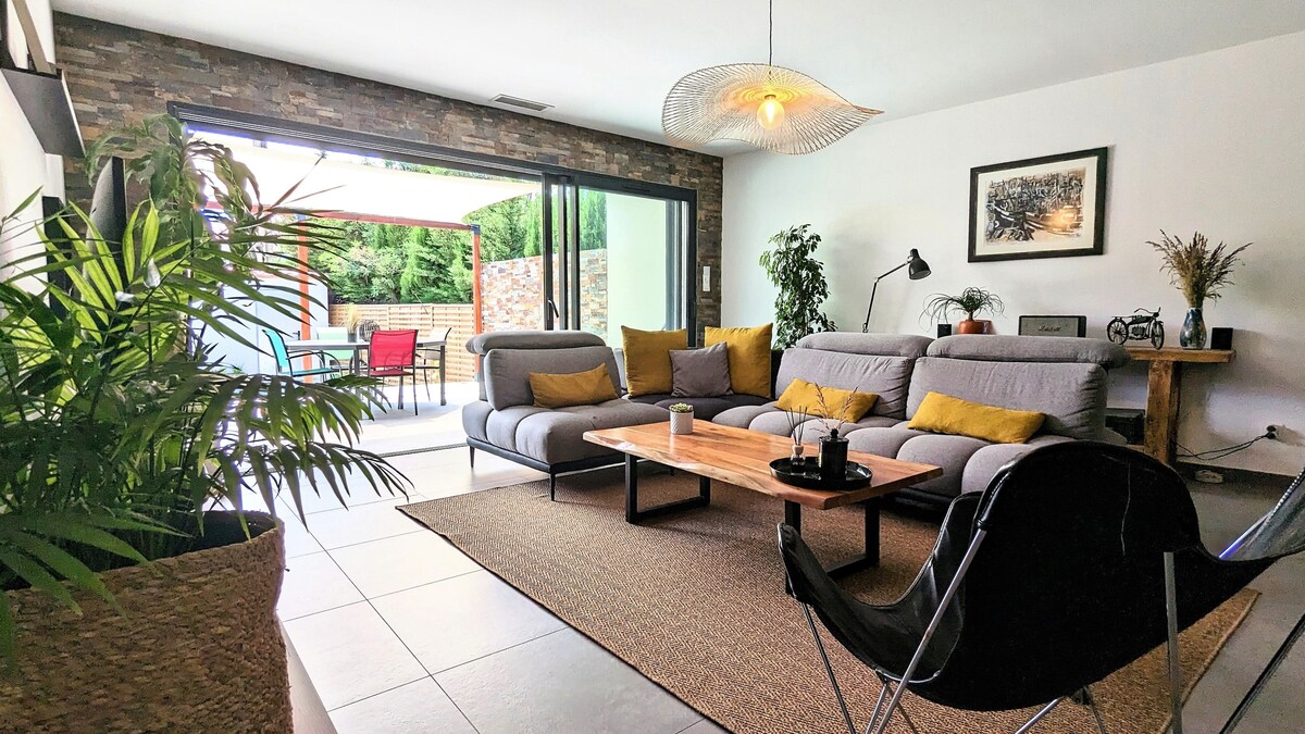 A spacious living area features a large sectional sofa with yellow accent pillows, complemented by a wooden coffee table. Floor-to-ceiling glass doors provide a view into the outdoor terrace, while plants add a touch of greenery to the room's natural aesthetic.
