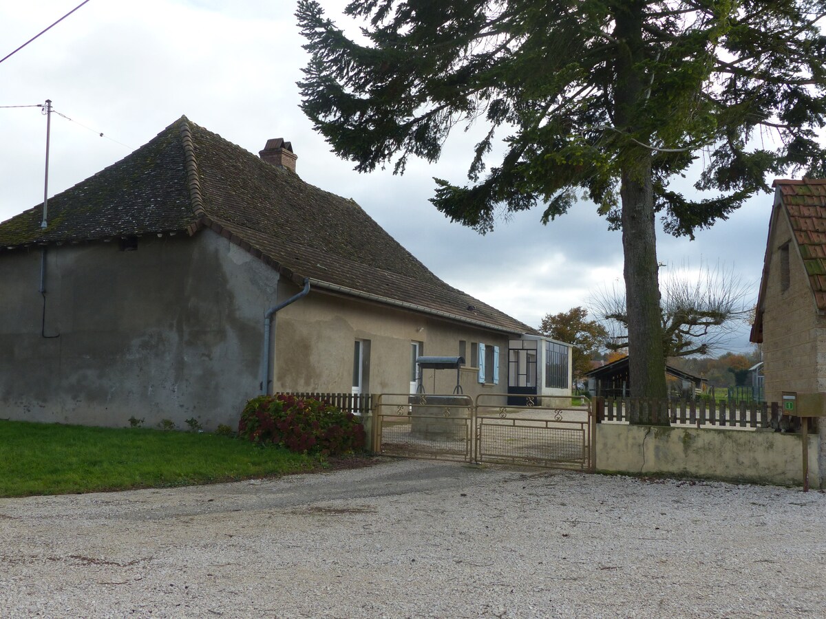 A country house with a sloped roof and a large, grassy front yard is visible. A gravel driveway leads to a fenced area, with distant trees and another building seen to the right. The surrounding landscape appears peaceful and open.